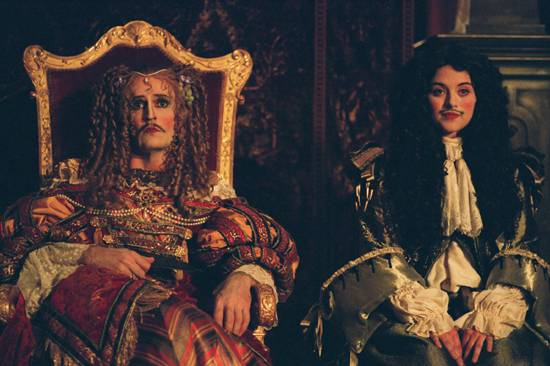 Maria and Ned both dressed as women and Royals, sitting on throne like chairs with unamused expressions.