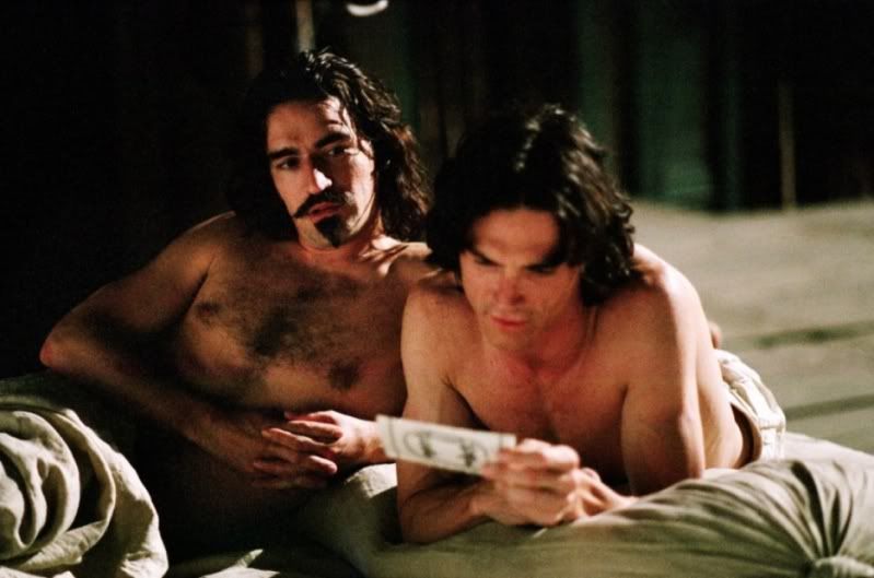 George looks over at Ned while he reads a paper. Both are laying in bed covered with bedsheets.