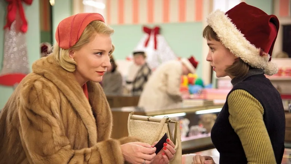 Carol apoyada contra el mostrador hablando con Therese en una tienda de dulces. Therese se ve nerviosa y Carol m&aacute;s confiada.