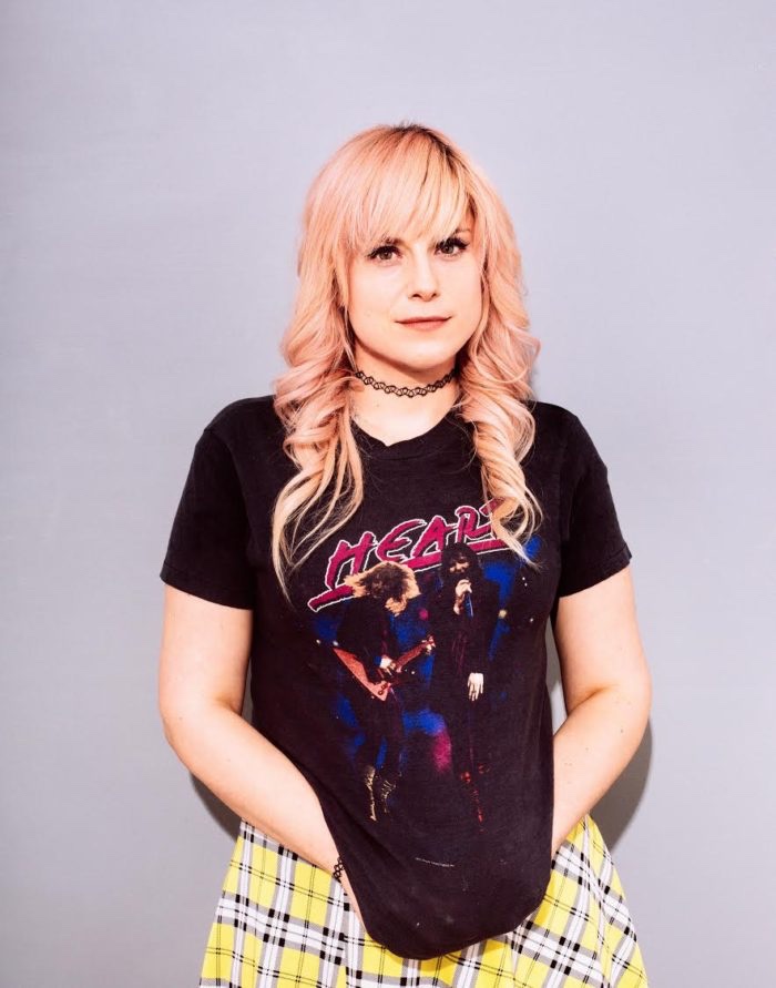 Lane More with dyed hair and a rocker shirt smiling with her hands in front.