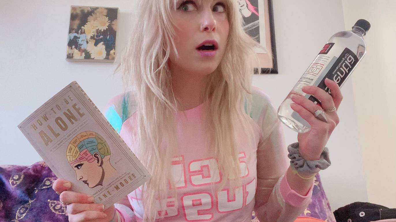 Lane Moore with a shocked expression holding her book in one hand and a water bottle in the other in her room.