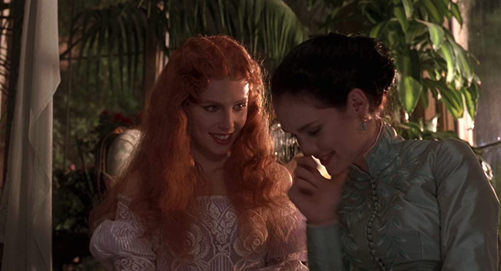 Lucy y Mina después de compartir un beso, riendo para sí mismas sentadas cerca de un jardín.