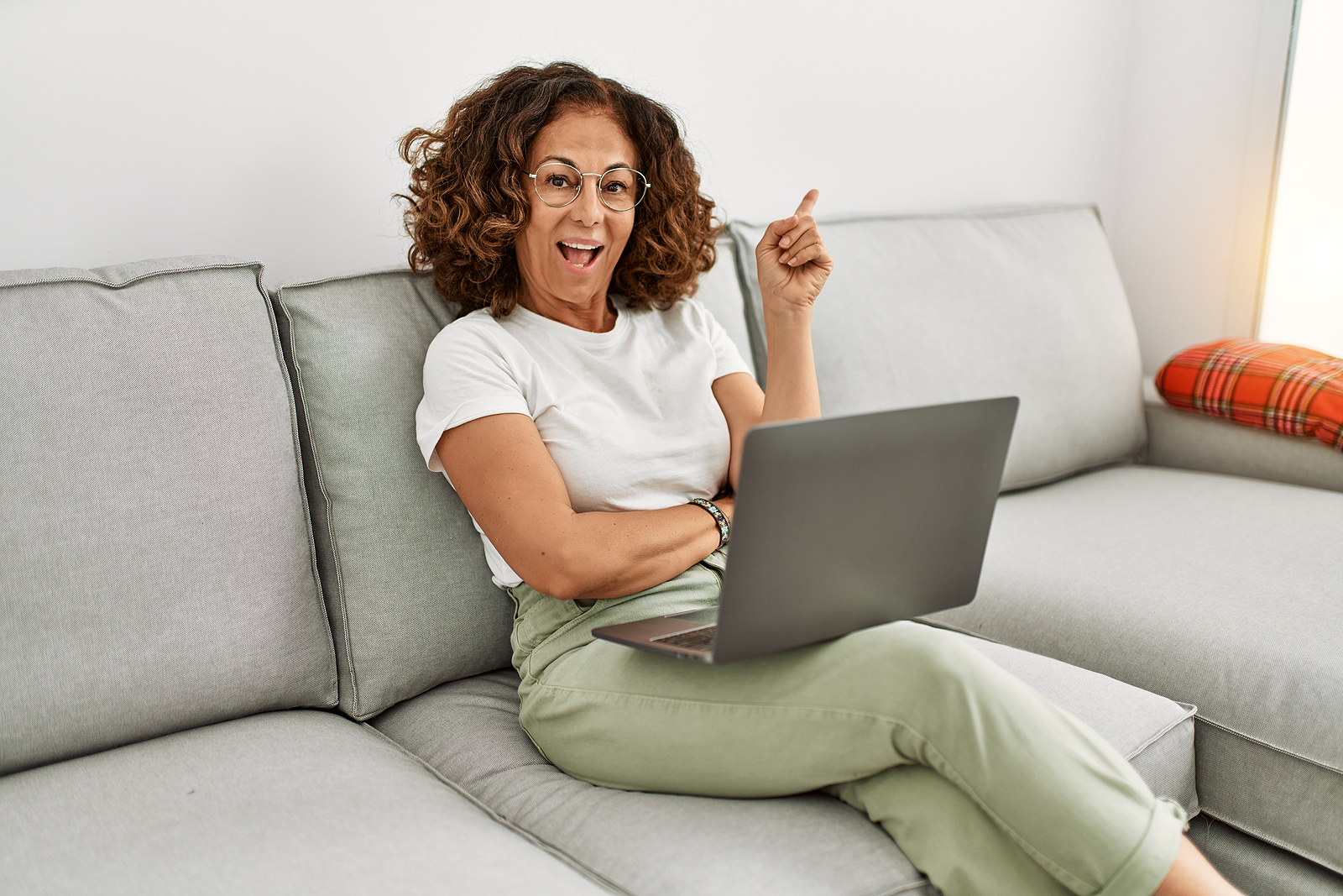 Una mujer latina de mediana edad sonr&iacute;e mientras trabaja en su laptop en su sof&aacute; con un dedo apuntando hacia afuera.