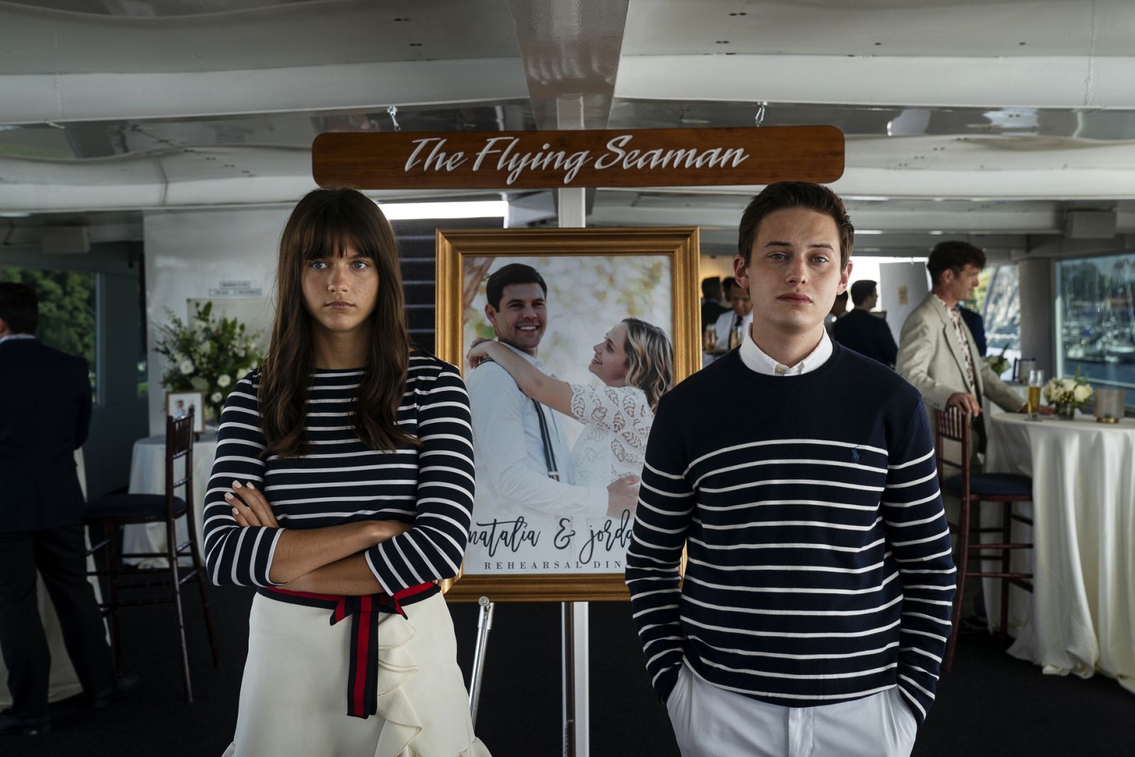 Naomi and Nathan wearing striped sweaters at a boat event with solemn expressions and hands in their pockets.