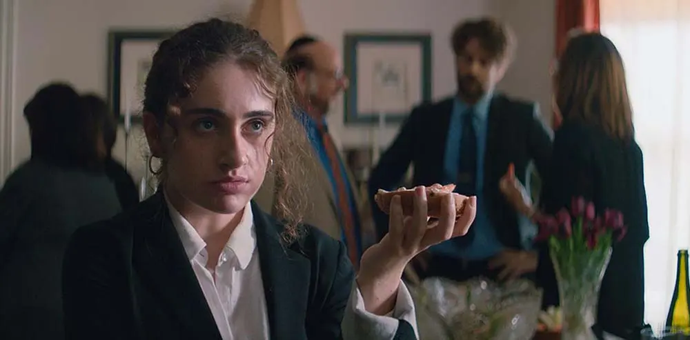 Danielle holding a bagel in one hand looking to the side while Jewish relative are in the background.