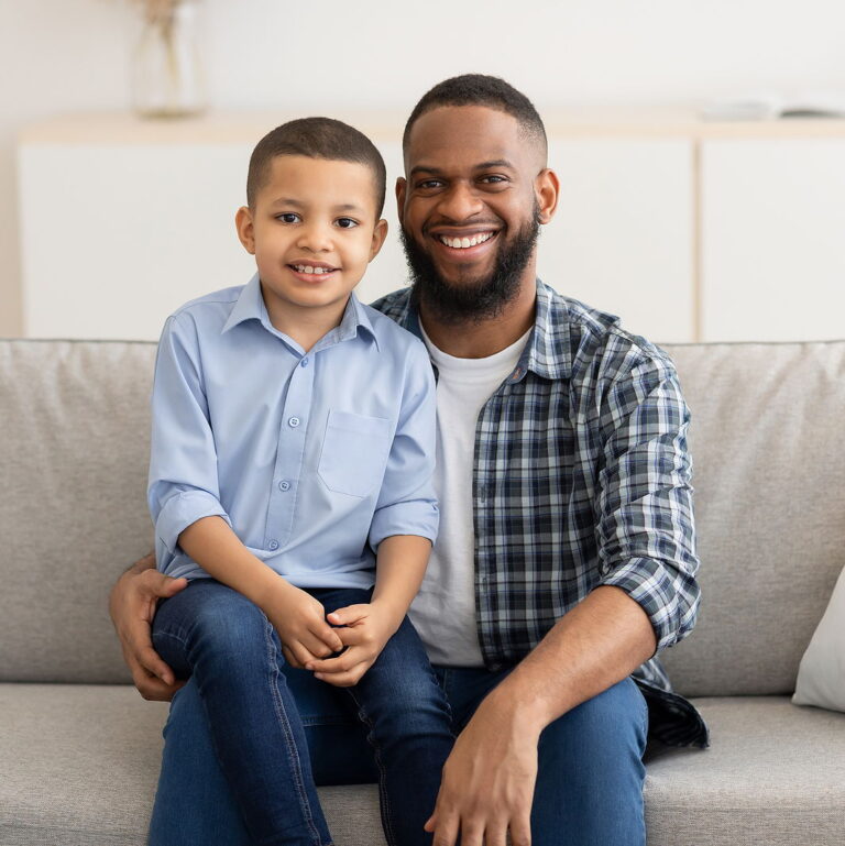 A black man sits on a couch with his young son on his lap. Both are smiling looking at the camera.