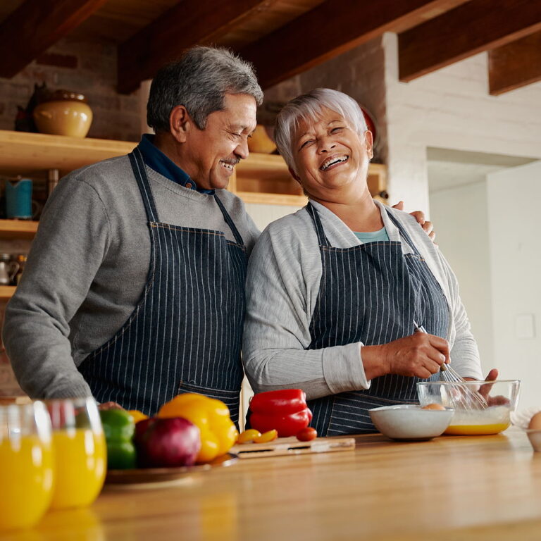 A bi racial married couple laugh together while making breakfast int heir kitchen. The husband has his hand on his wife's shoulder.