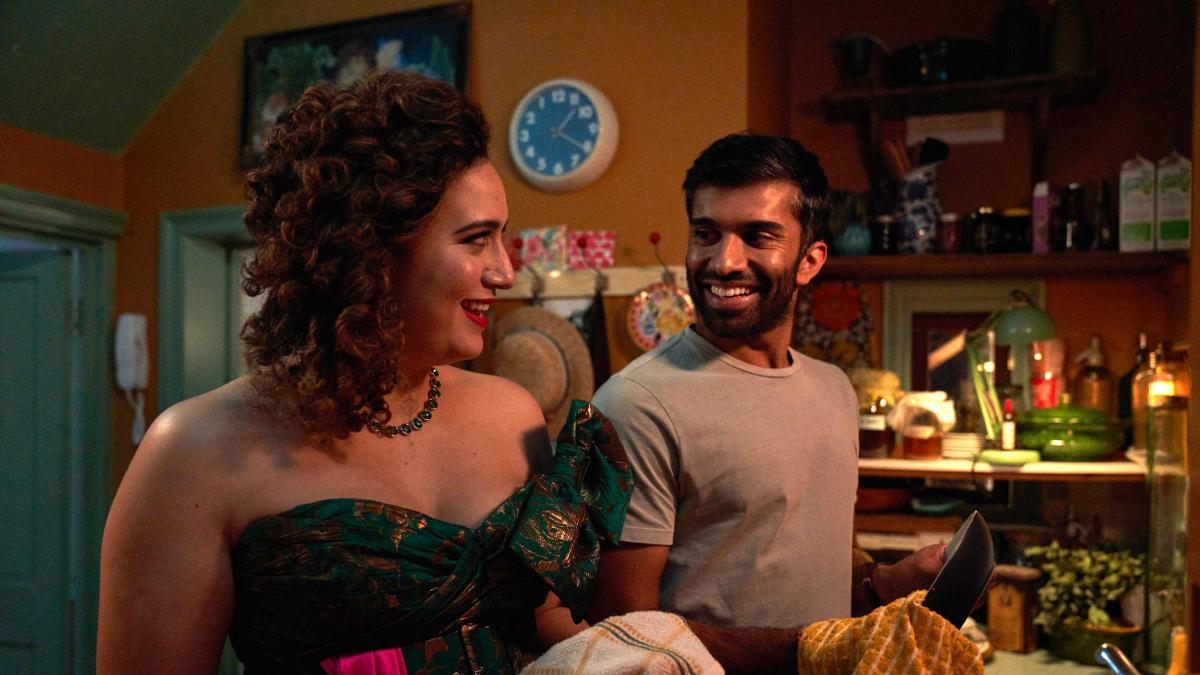 Jessie and Tom talking and smiling while they wash dishes together. Jessie has on a dress while tom a t shirt.