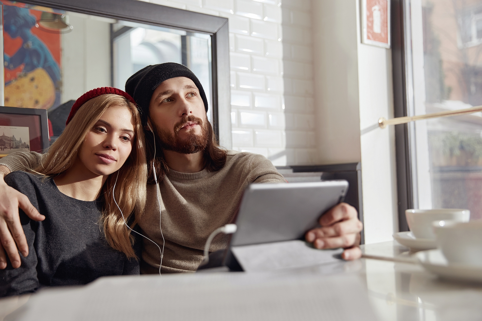 Joven pareja europea pensativa con auriculares viendo algo en una tableta digital en un caf&eacute; moderno.