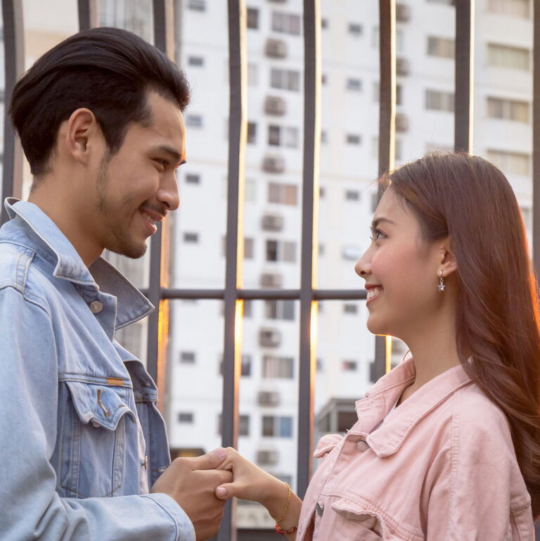 A young asian man and woman stand facing eachother. They are holding onto a hand and smiling.