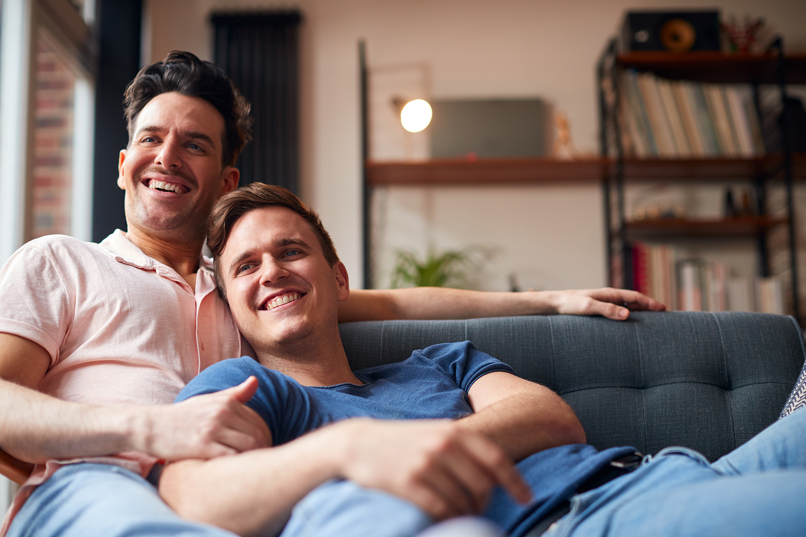 Una atractiva pareja gay est&aacute; abrazada en el sof&aacute; sonriendo viendo la televisi&oacute;n.