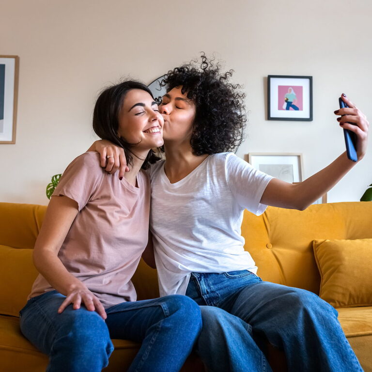 A young lesbian couple smiling sitting on their sofa taking a selfie together while sharing a kiss