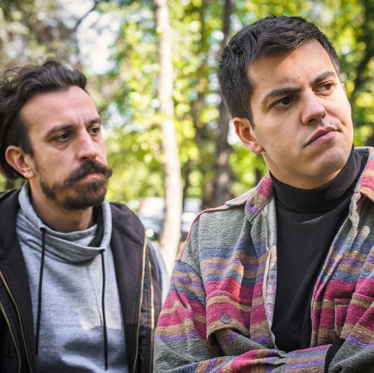 Two men in outdoor clothing look away from one another with one annoyed at the other.