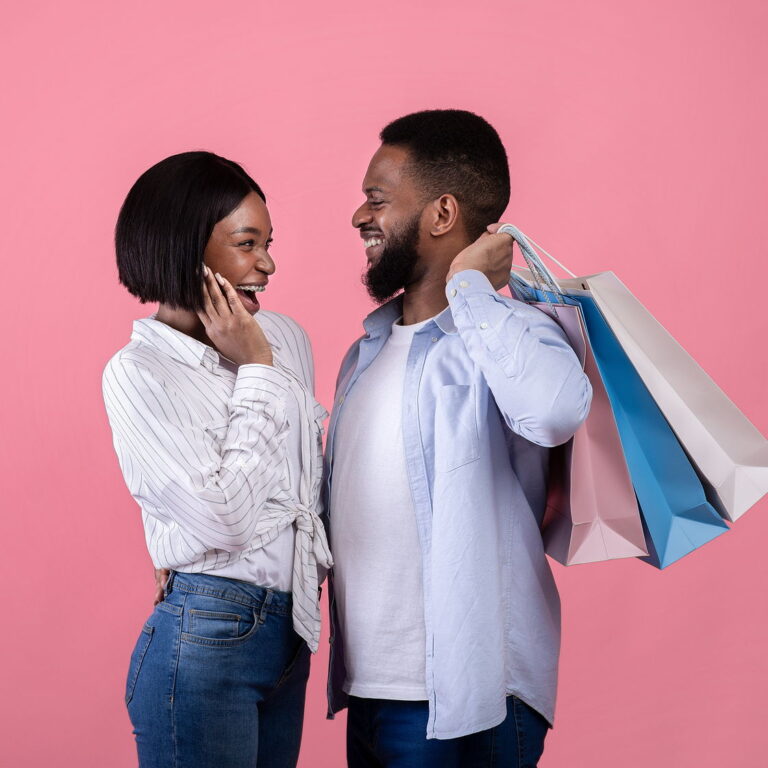 A young black couple smiling facing eachother. She has a surprised expression while he smiles holding shopping bags behind his back.