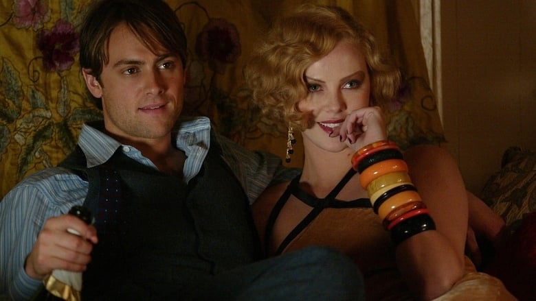 Guy and Gilda laying against a bed together laughing looking at someone out of frame. Guy holds a bottle of champagne.