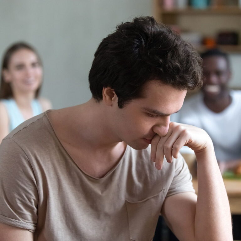 A man contemplates while his hand is reclining on his head. His friends are behind him smiling while looking in his direction.
