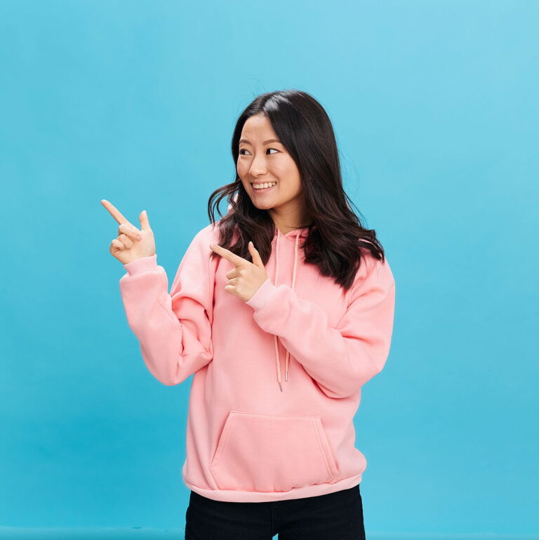 Overjoyed cute Asian student young lady in pink hoodie sweatshirt point finger to someone out of frame.