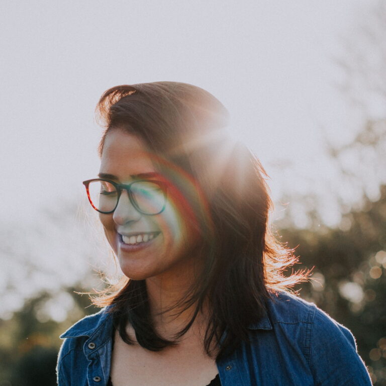 A woman with glasses looks down while outdoors. The sun is setting behind her.