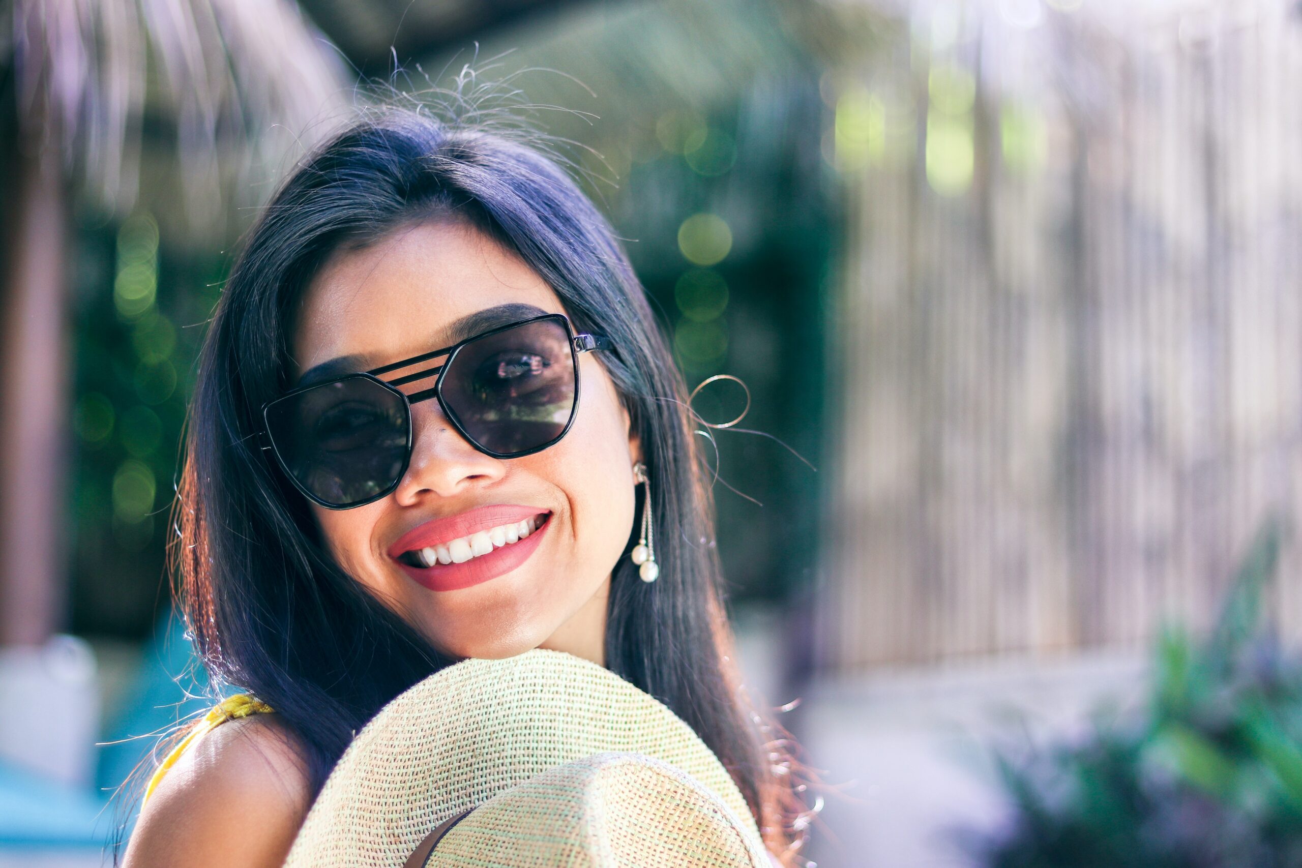 A latina woman smiles looking at the camera, wearing sunglasses and outdoors.