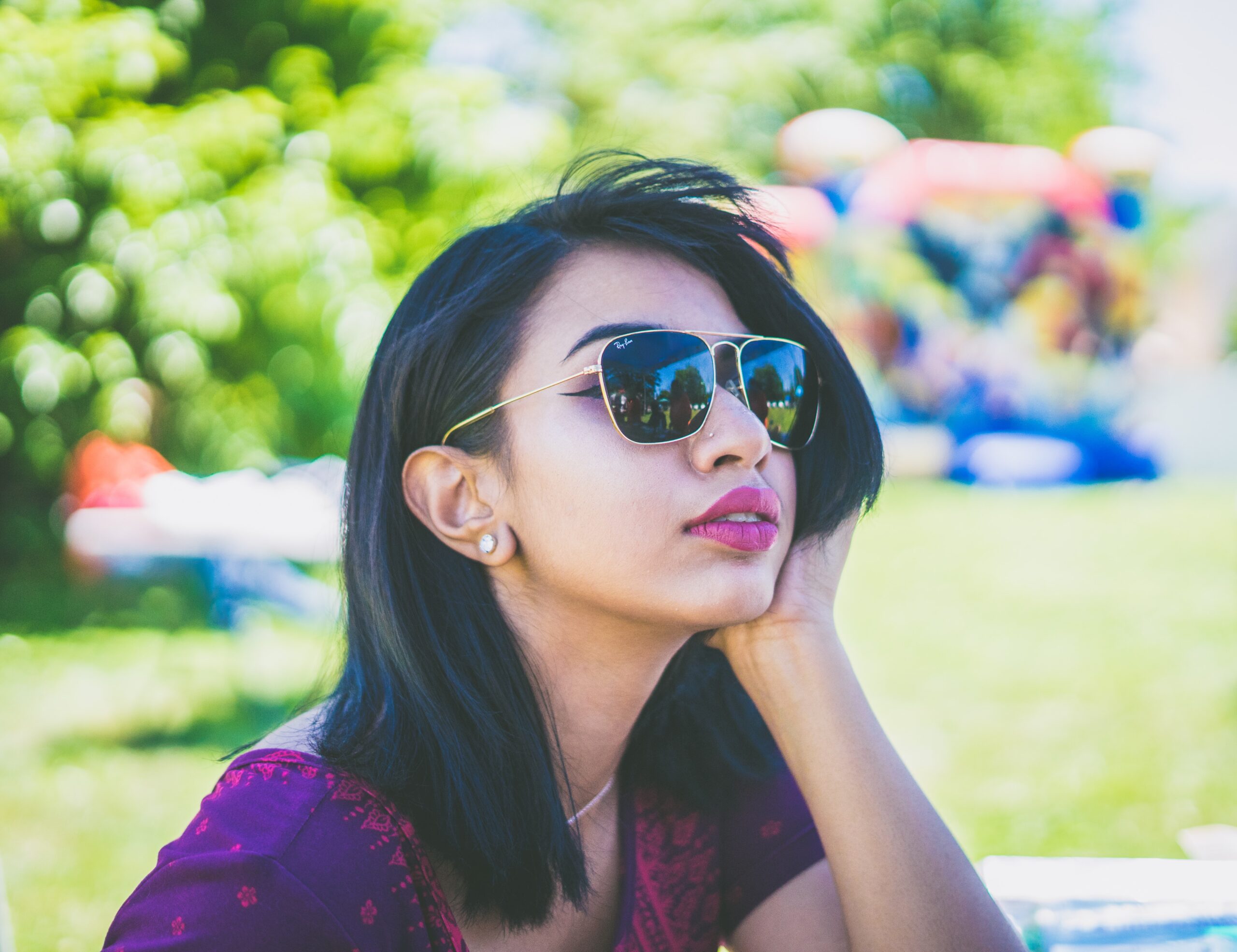 A woman with dark glasses sits on a bench looking away, she has a hand on her cheek.