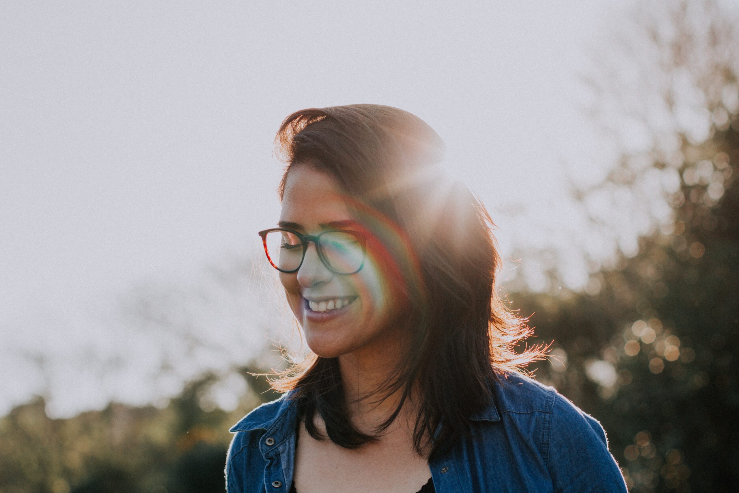 A woman with glasses looks down while outdoors. The sun is setting behind her.