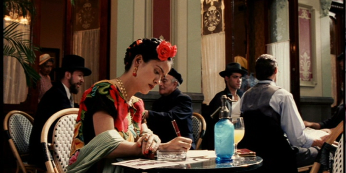 Frida sitting at a cafe, writing on a paper with pen.There are only men around her.