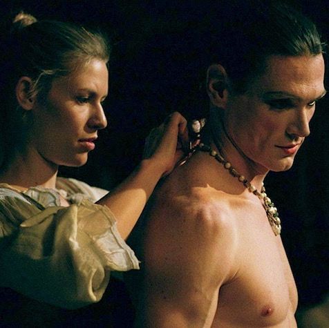 Maria helps a shirtless Ned remove part of a necklace and costume from behind him.