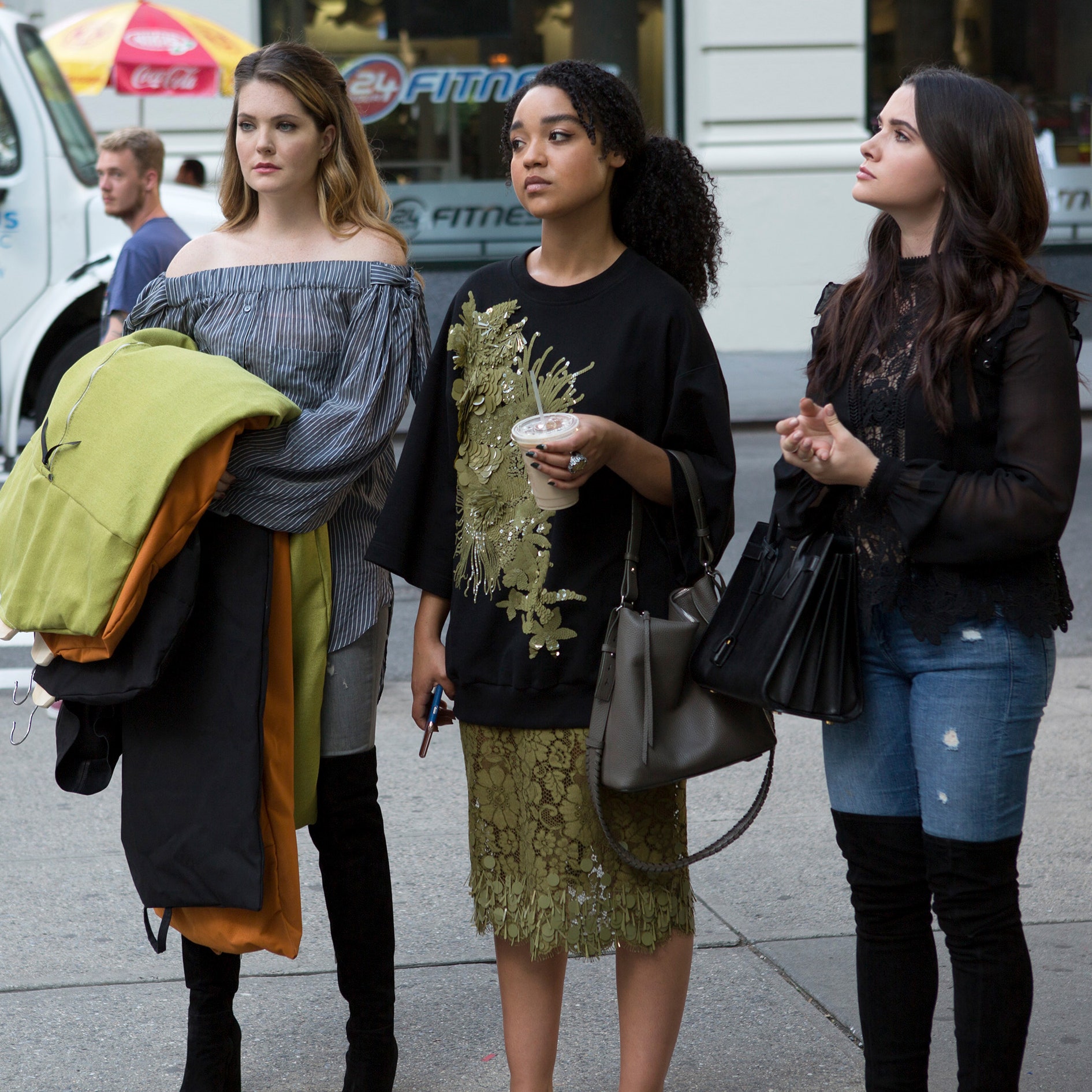 Jane, Sutton y Kat looking somewhat annoyed to the side, all standing in the street holding coffee, clothes, and their purses.