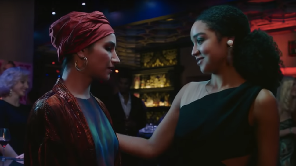 Kat reaching out to Adena who is wearing a headscarf, both are in a bar with dim lights.