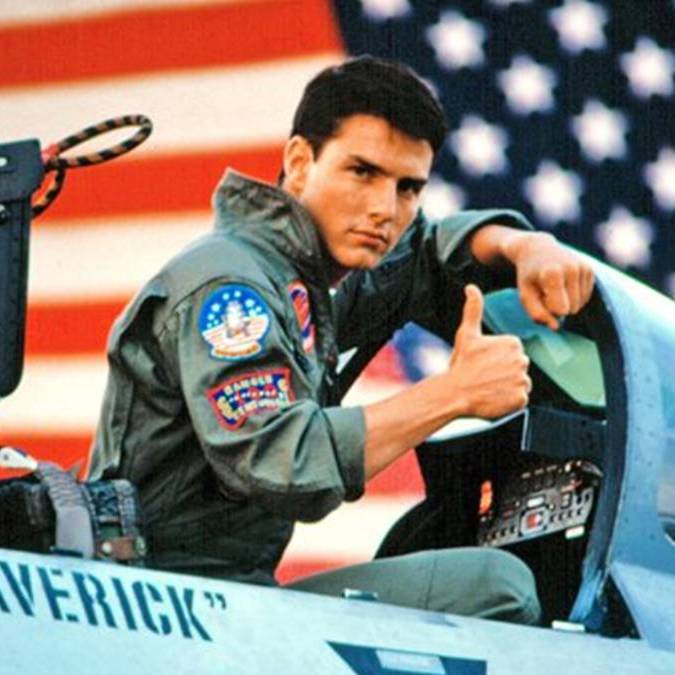Maverick giving the thumbs up sitting inside a one of his fighter planes with a large american flag behind him.