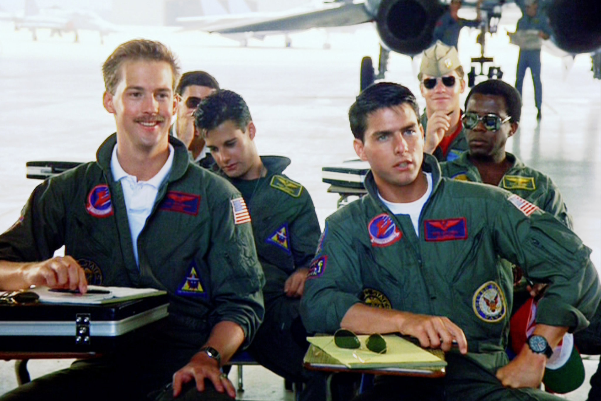 Maverick y Goose sentados en escritorios con otros en su uniforme, escuchando a un orador. Goose est&aacute; sonriendo y Maverick tiene una mirada seria.