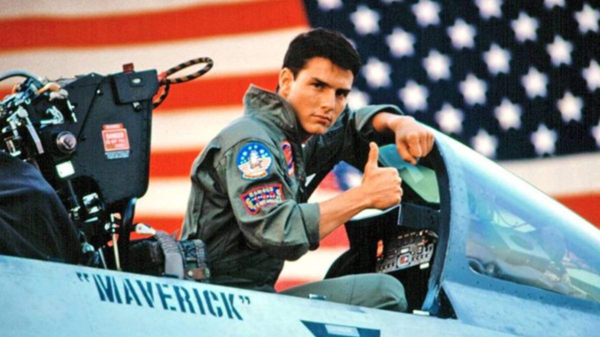 Maverick con el dedo afuera en afirmacion sentado dentro de uno de sus aviones de combate con una gran bandera americana detr&aacute;s de &eacute;l.