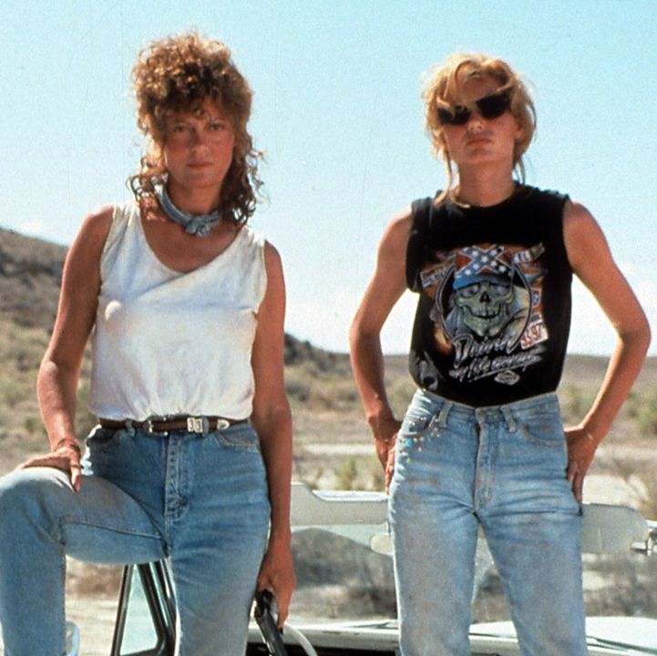 Thelma and Louise posing looking cool. Louise holding a handgun. Standing in front of their parked car on the road.