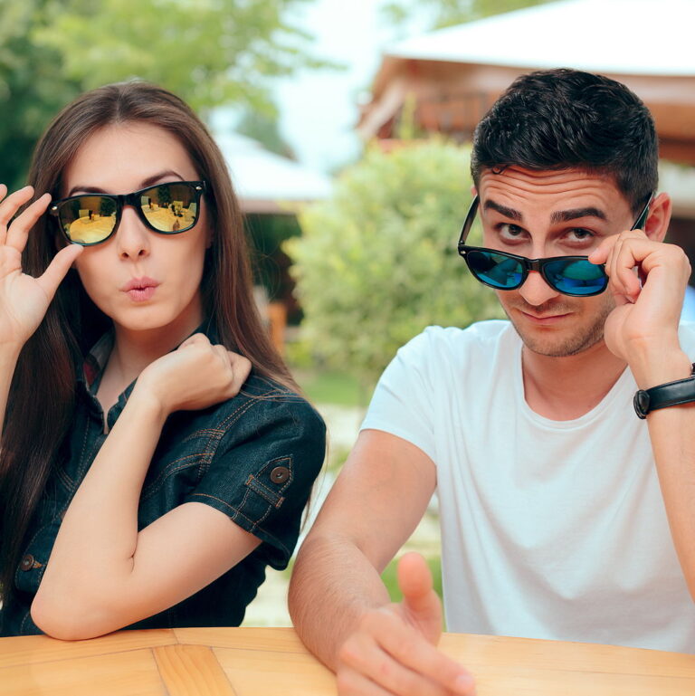 An attractive man and woman sitting together with sunglasses and a judgy expression on their faces looking at the camera.