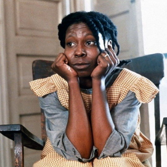 Celie with both her hands on her lap holding her face looking at a distance sitting on a chair.