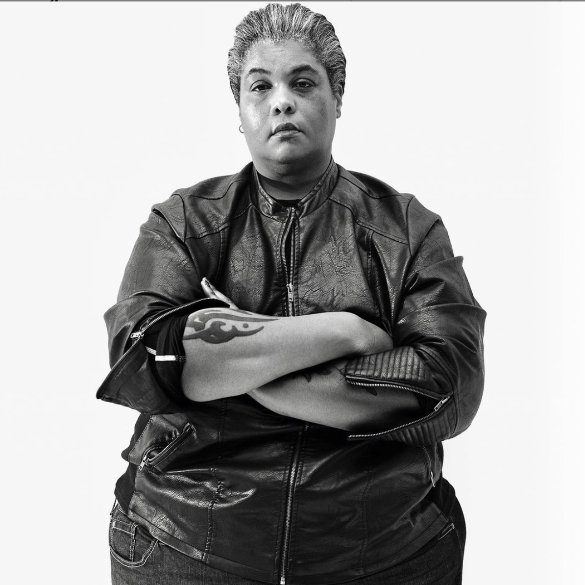 Black and white image of Roxane standing confidently with her arms crossed wearing a leather jacket. Her tattoo on her arm is exposed.