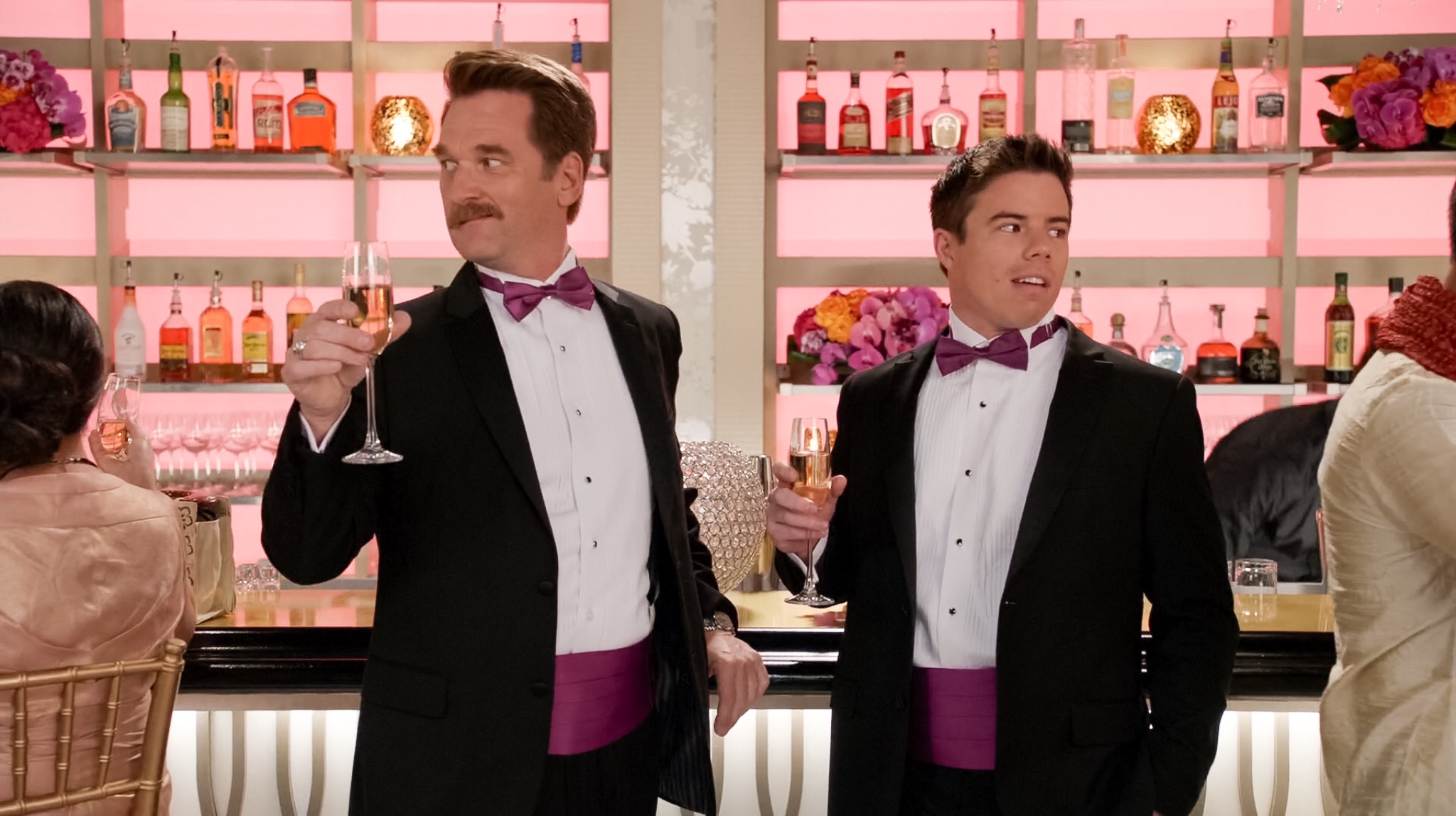 Daryll and Josh wearing tuxedos for a wedding standing at the bar with a drink in hand looking in opposite directions.