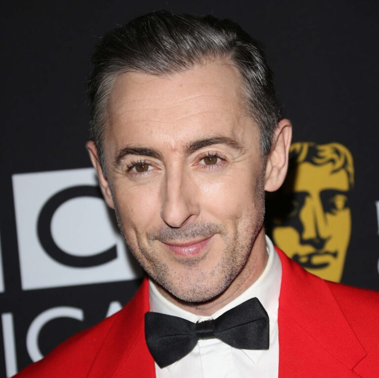 Alan Cumming at a carpet event wearing a red suit and bowtie smiling to the camera.