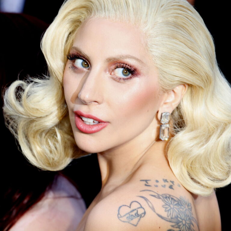 Lady Gaga at a carpet event smiling looking behind her shoulder. Her dress shows off back tattoo.