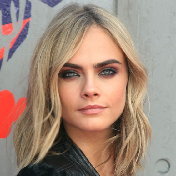 Cara Delevingne at an event smiling and looking to the side. She is dressed up and wearing a silver necklace.