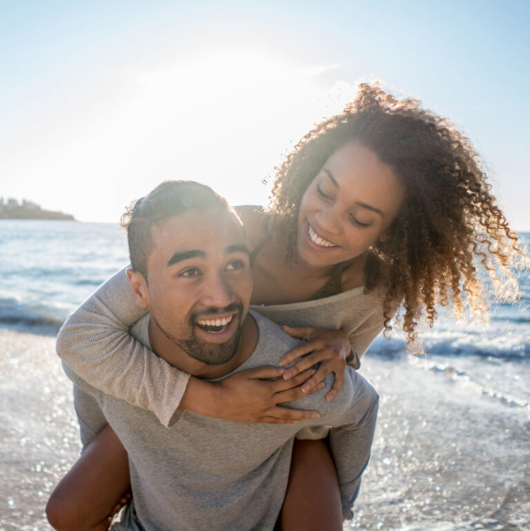 A multi ethnic couple smiling together at the beach. The woman is on the mans back holding onto him.
