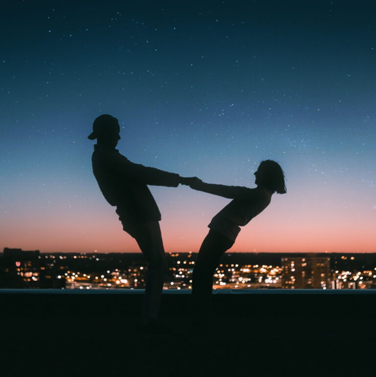 A the silouhette of a man and a woman holding both hands together and leaning back, during dusk.