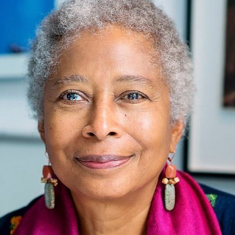 An older Alice Walker smiling warmly to the camera. She is in front of large pictures on the wall.