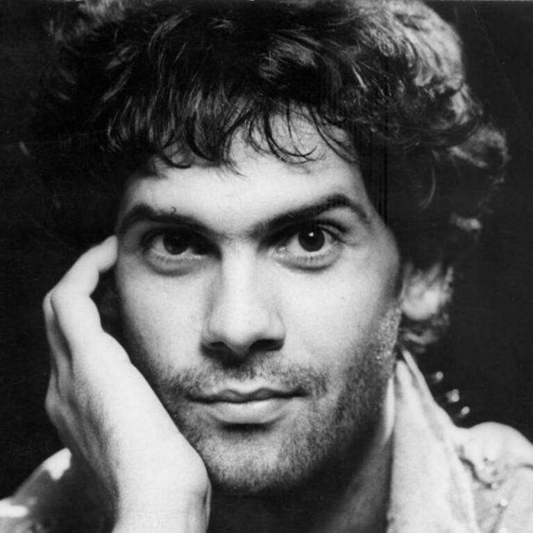 Black and white image of Cazuza with a hand on his cheek smiling to the camera.