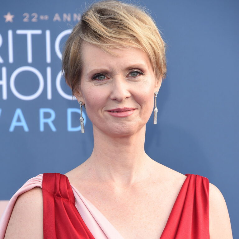 Cynthia Nixon at a carpet event wearing a dress and smiling.