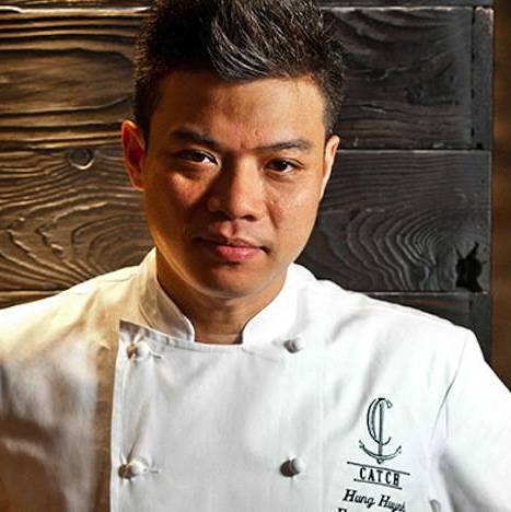 Hung smiling wearing a chefs apron and white coat.