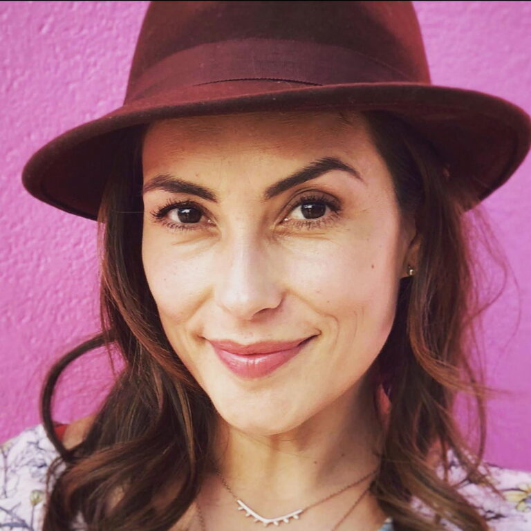 Iyari wearing a hat and smiling against a bright colored wall.