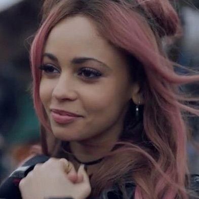 Toni Topaz smiling and holding her hand close to her chest. She has her signature pink hair color.
