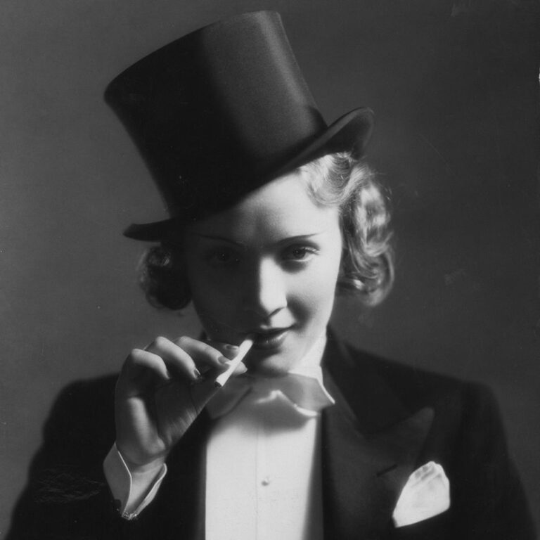 Black and white image of Marlene Dietrich wearing a tux and top hat with a cigarette in one hand.