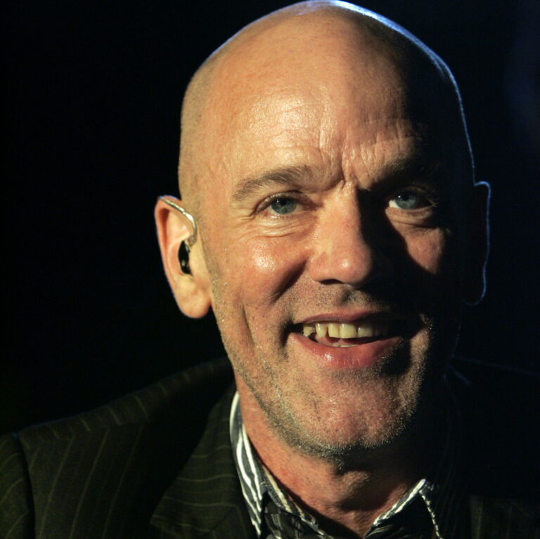 Michael Stipe smiling against a dark background wearing a suit.